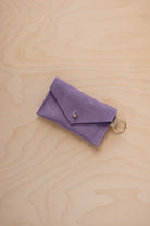 A small lavender coloured suede envelope-style keyring pouch with a gold button, resting on a light wood grain surface.