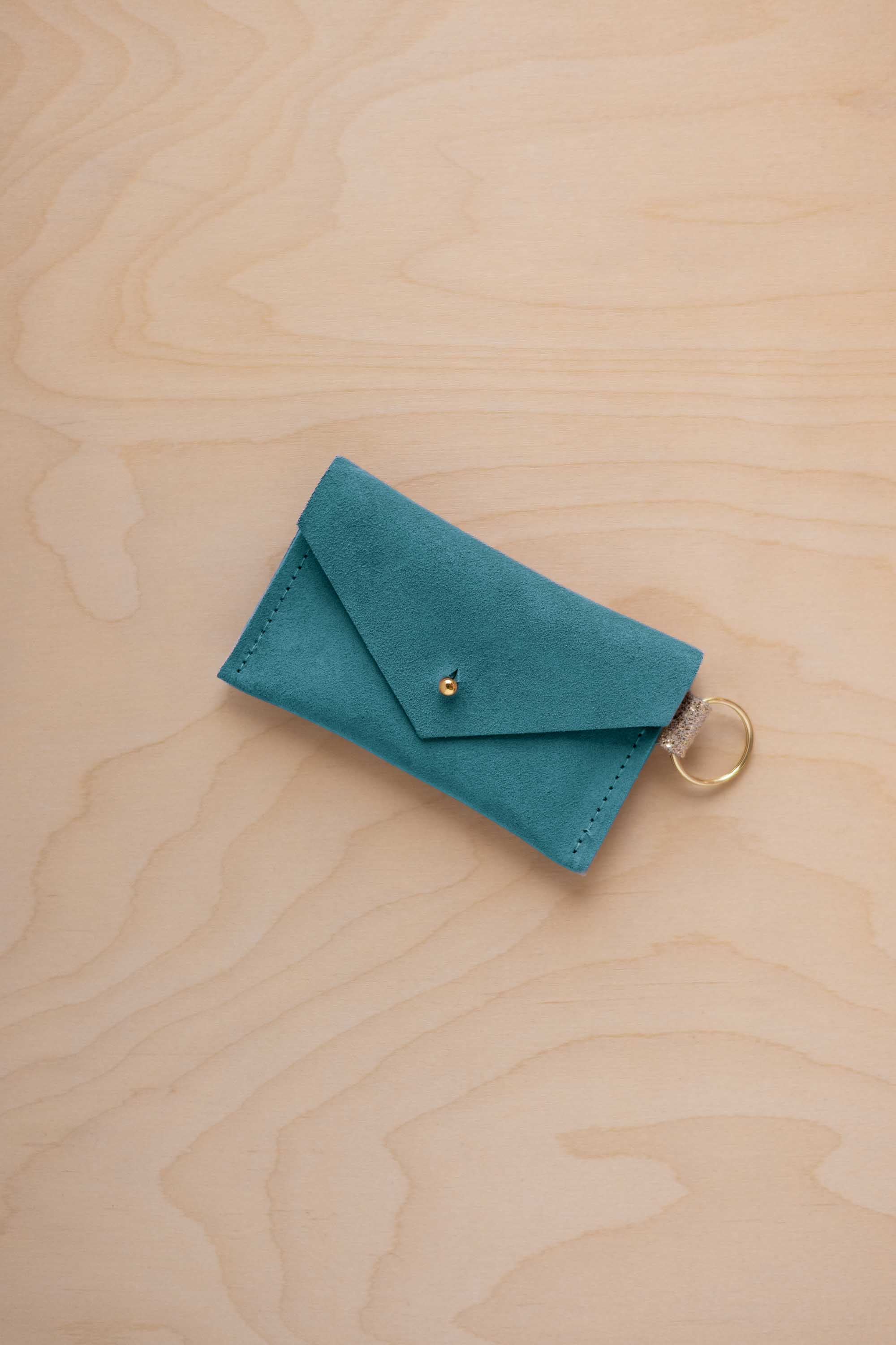 A small marine teal coloured suede envelope-style keyring pouch with a gold button, resting on a light wood grain surface.