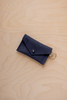 A small midnight navy coloured suede envelope-style keyring pouch with a gold button, resting on a light wood grain surface.