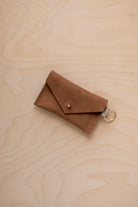 A small mocha brown coloured suede envelope-style keyring pouch with a gold button, resting on a light wood grain surface.