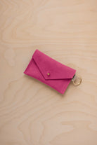 A small raspberry coloured suede envelope-style keyring pouch with a gold button, resting on a light wood grain surface.