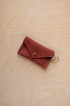 A small rust red coloured suede envelope-style keyring pouch with a gold button, resting on a light wood grain surface.