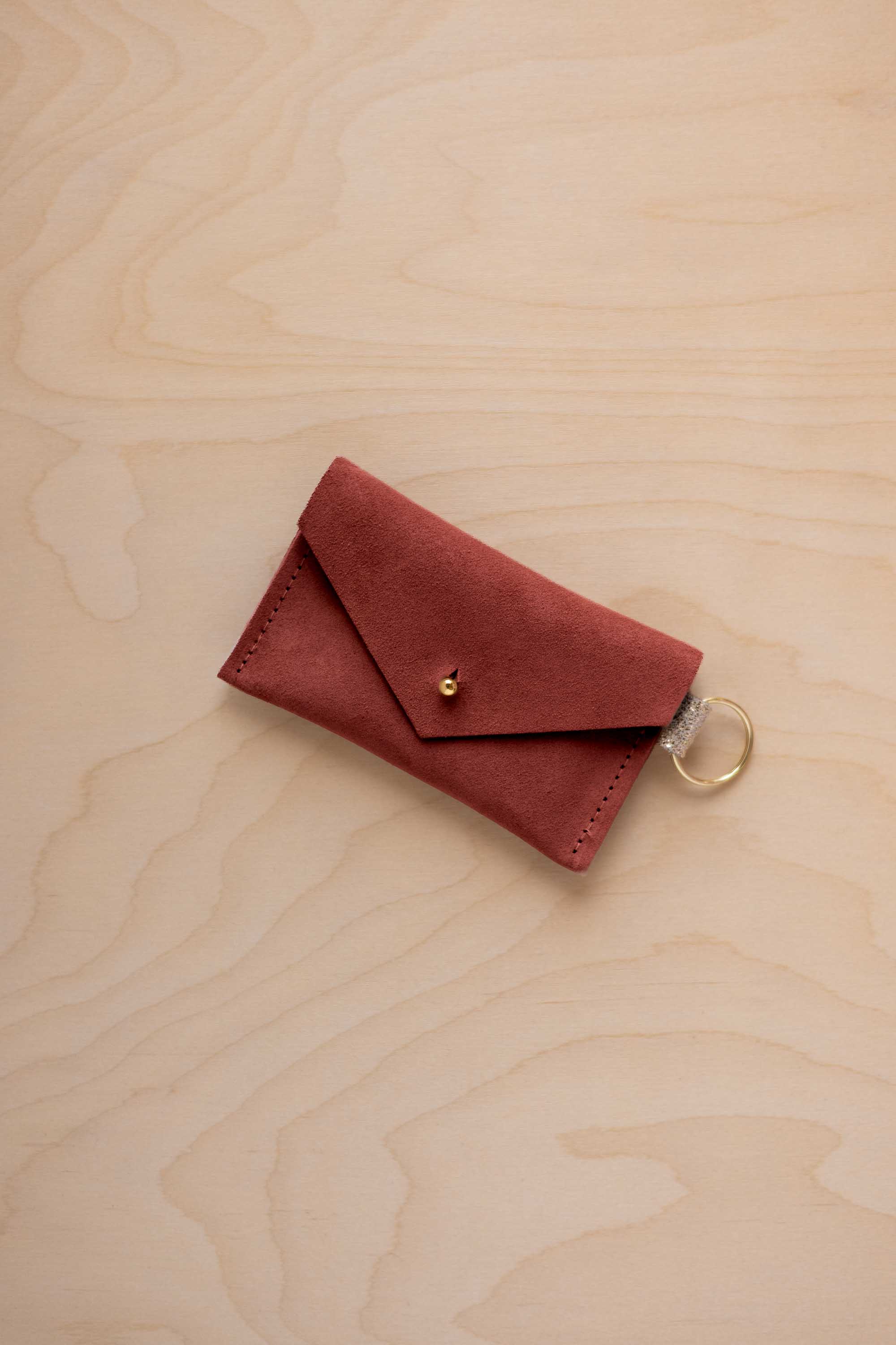 A small rust red coloured suede envelope-style keyring pouch with a gold button, resting on a light wood grain surface.