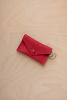 A small scarlet red coloured suede envelope-style keyring pouch with a gold button, resting on a light wood grain surface.