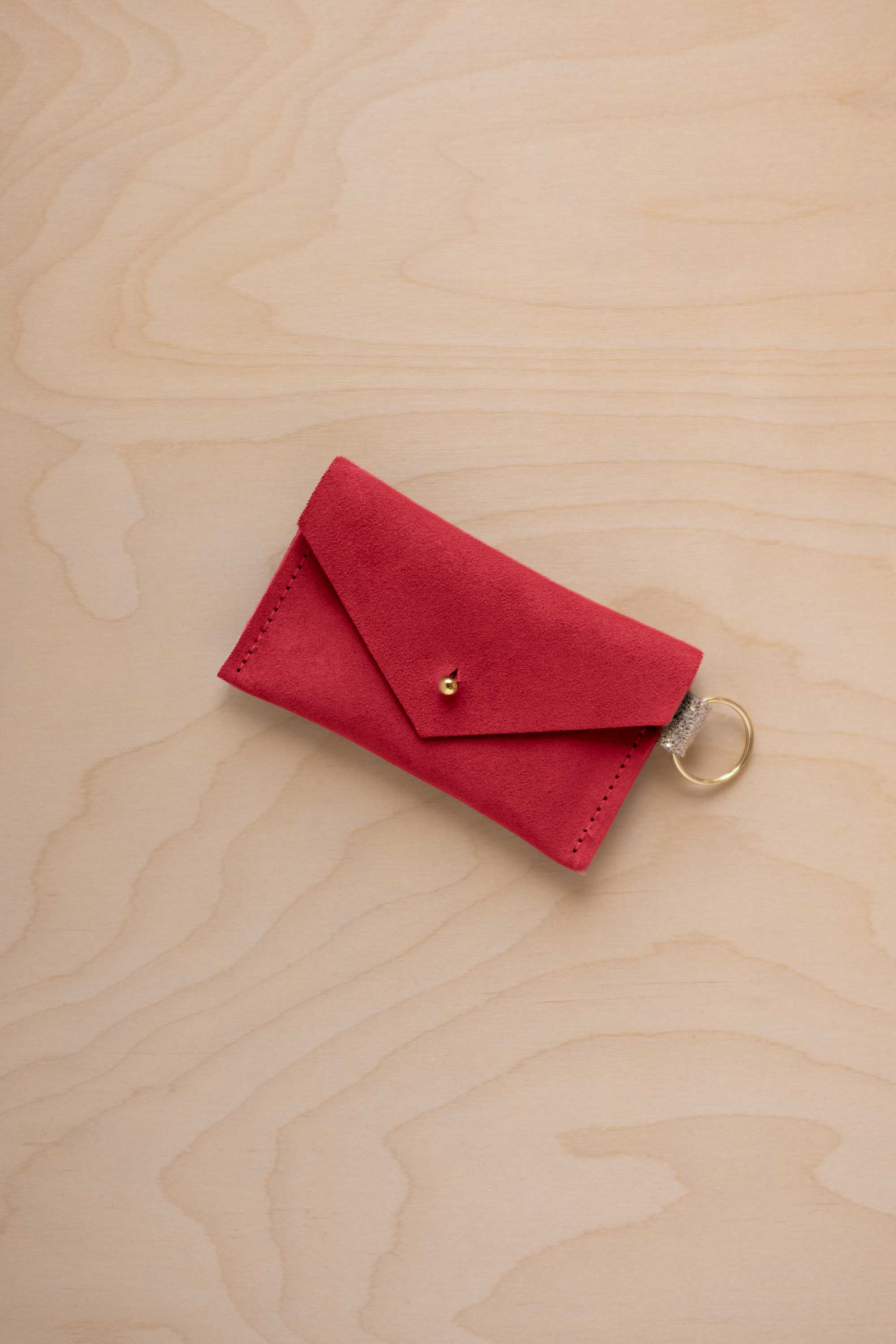 A small scarlet red coloured suede envelope-style keyring pouch with a gold button, resting on a light wood grain surface.