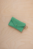 A small seafoam green coloured suede envelope-style keyring pouch with a gold button, resting on a light wood grain surface.