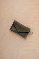 A small soft olive green coloured suede envelope-style keyring pouch with a gold button, resting on a light wood grain surface.