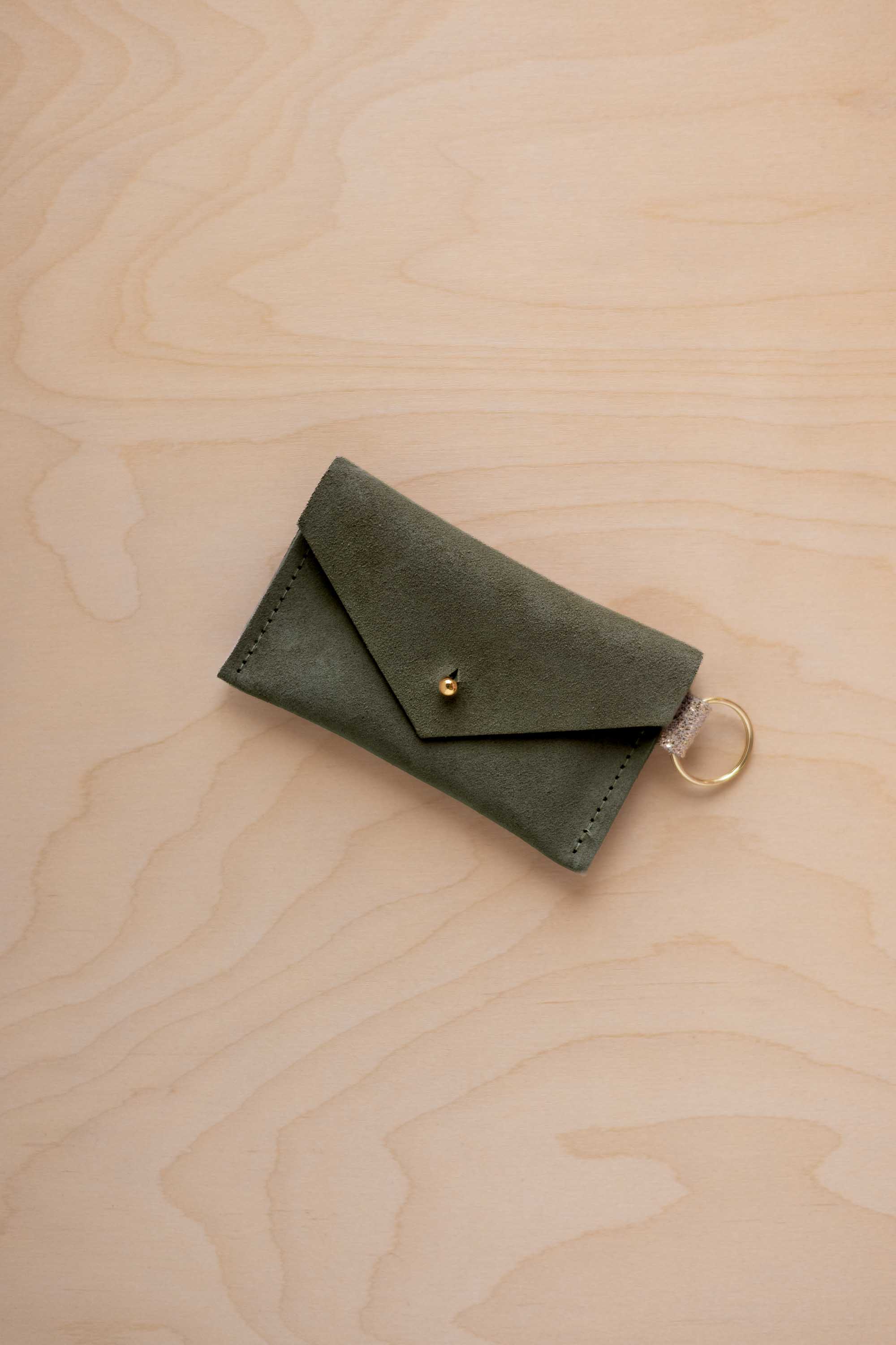 A small soft olive green coloured suede envelope-style keyring pouch with a gold button, resting on a light wood grain surface.