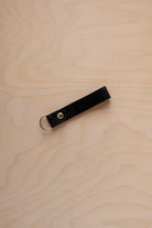 A black suede leather keychain with a gold metal ring and rivet, displayed diagonally on a light wood grain background.