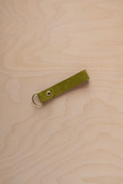 A chartreuse lime green coloured suede leather keychain with a gold metal ring and rivet, displayed diagonally on a light wood grain background.