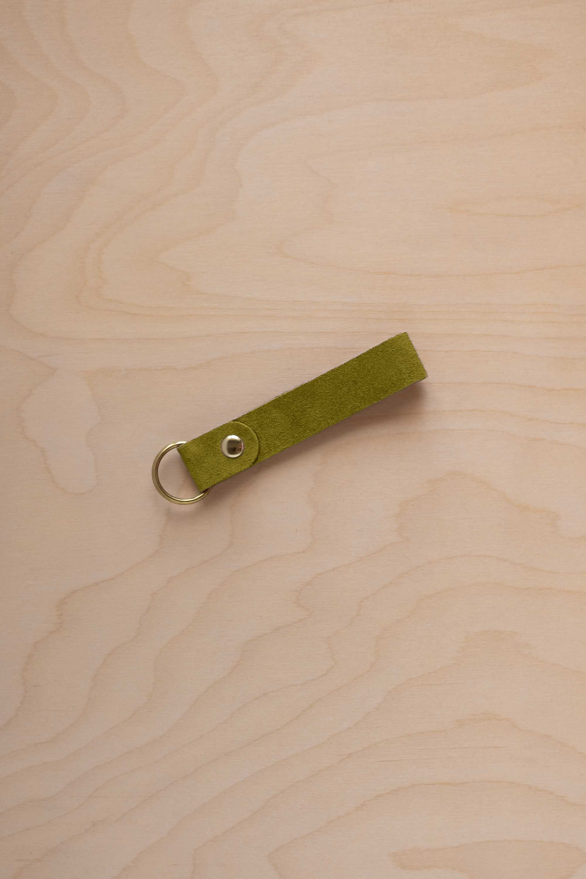 A chartreuse lime green coloured suede leather keychain with a gold metal ring and rivet, displayed diagonally on a light wood grain background.