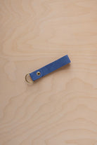 A cobalt blue suede leather keychain with a gold metal ring and rivet, displayed diagonally on a light wood grain background.