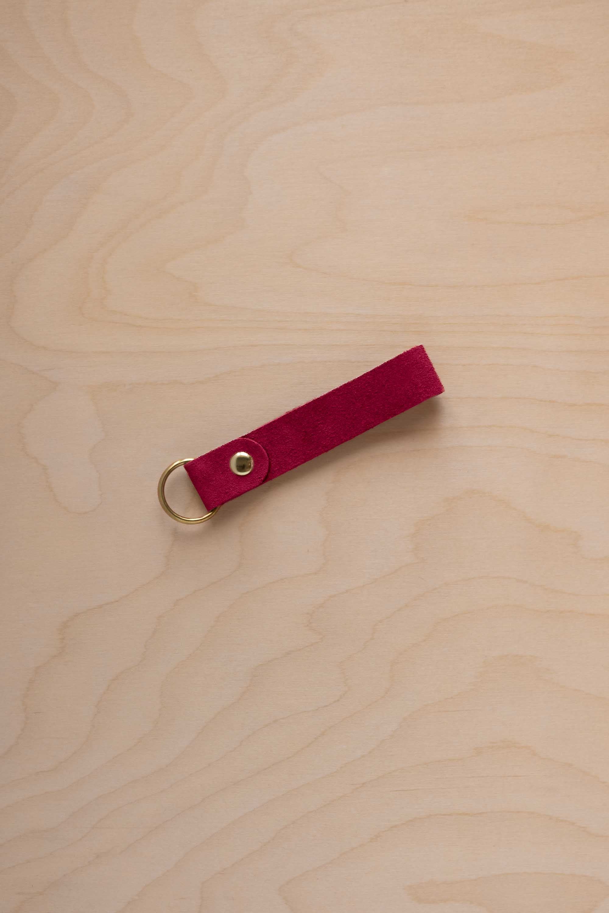 A crimson red suede leather keychain with a gold metal ring and rivet, displayed diagonally on a light wood grain background.