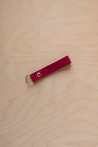 A crimson red suede leather keychain with a gold metal ring and rivet, displayed diagonally on a light wood grain background.