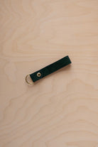 A deep forest green suede leather keychain with a gold metal ring and rivet, displayed diagonally on a light wood grain background.