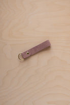 A dusty mauve suede leather keychain with a gold metal ring and rivet, displayed diagonally on a light wood grain background.