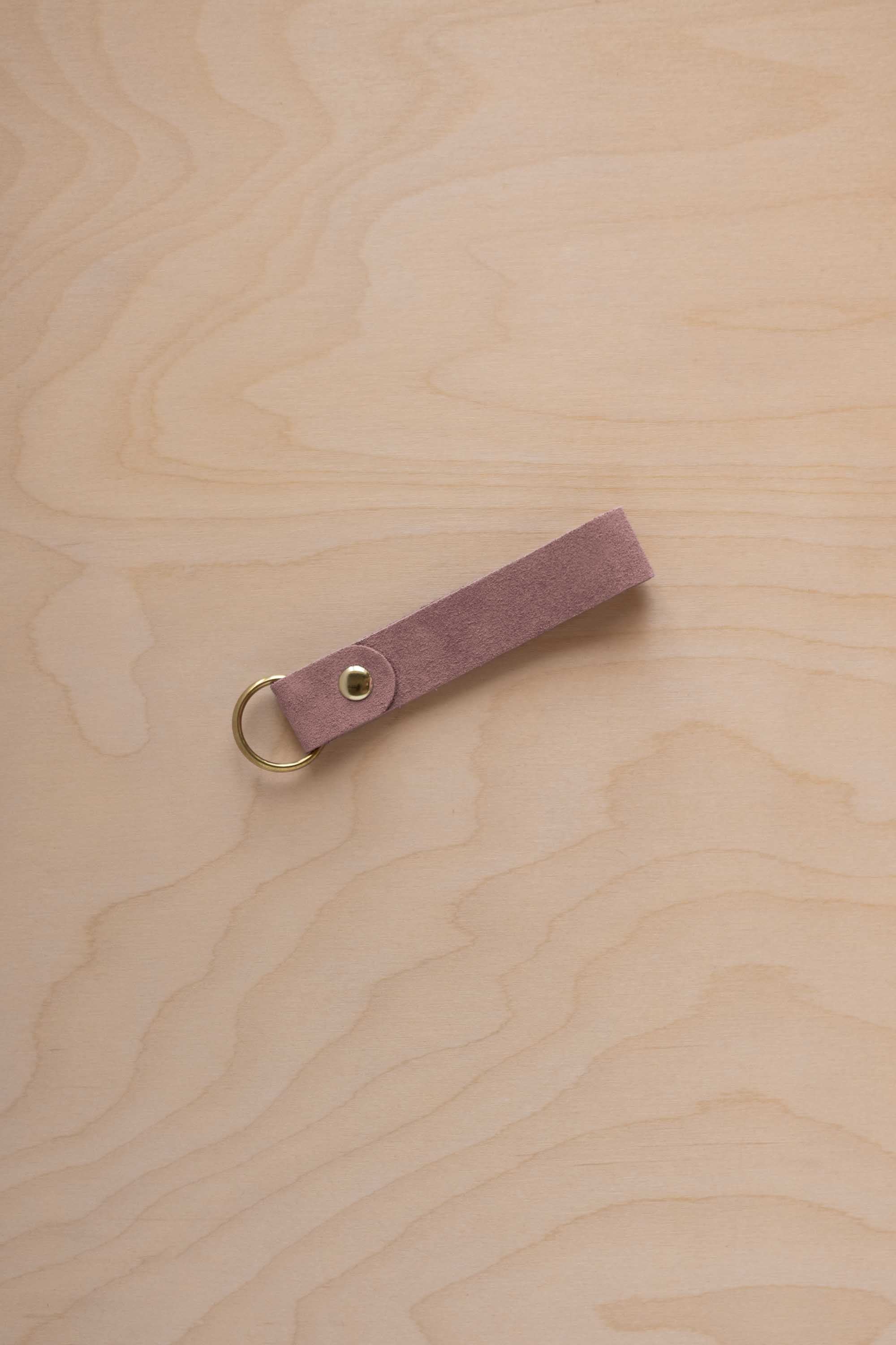 A dusty mauve suede leather keychain with a gold metal ring and rivet, displayed diagonally on a light wood grain background.