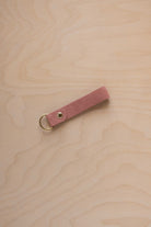 A dusty rose pale pink coloured suede leather keychain with a gold metal ring and rivet, displayed diagonally on a light wood grain background.