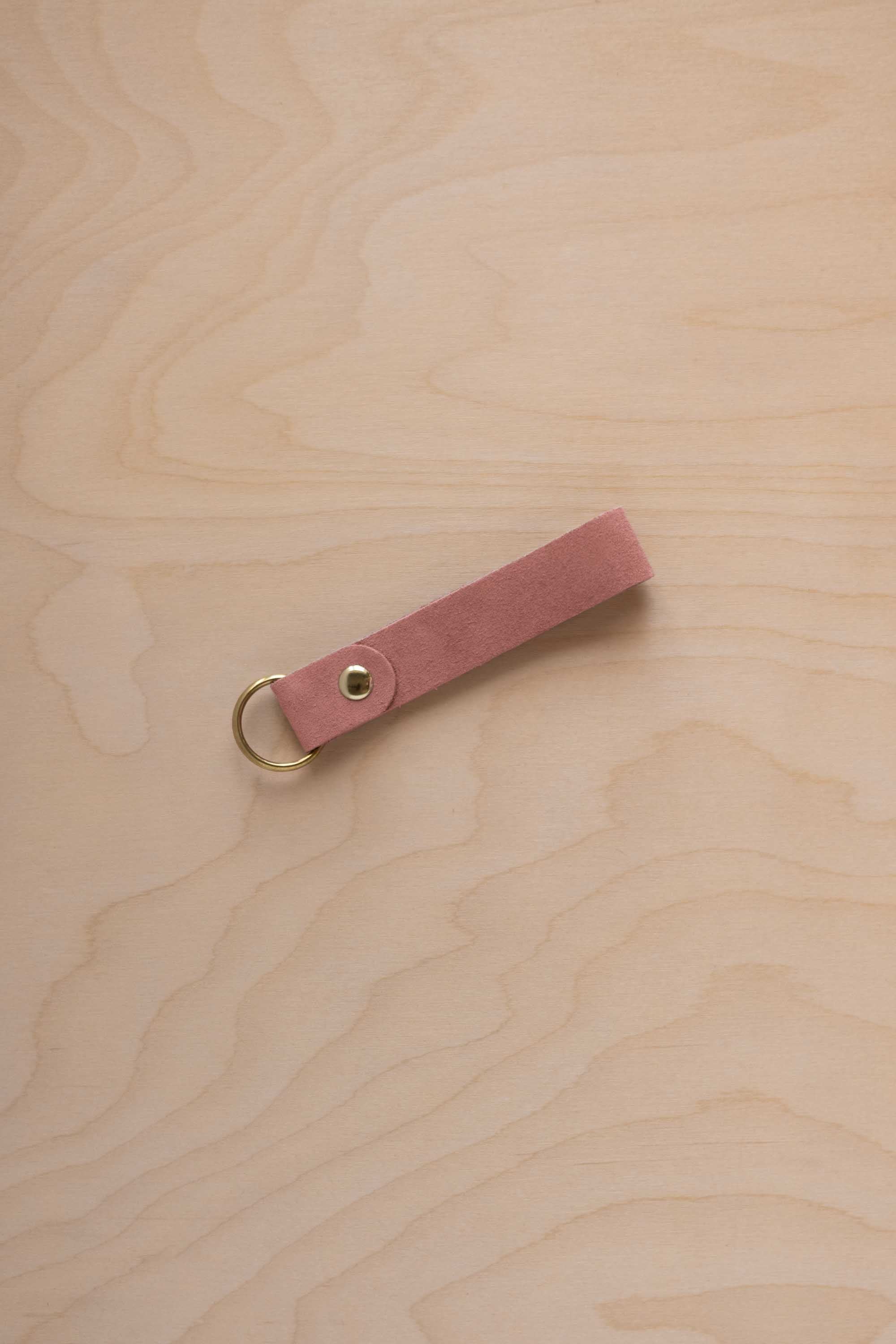 A dusty rose pale pink coloured suede leather keychain with a gold metal ring and rivet, displayed diagonally on a light wood grain background.