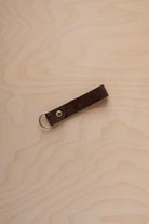 An espresso brown suede leather keychain with a gold metal ring and rivet, displayed diagonally on a light wood grain background.