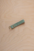 A eucalyptus mint green coloured suede leather keychain with a gold metal ring and rivet, displayed diagonally on a light wood grain background.