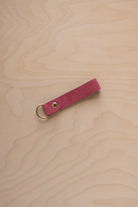 A faded rose pink coloured suede leather keychain with a gold metal ring and rivet, displayed diagonally on a light wood grain background.