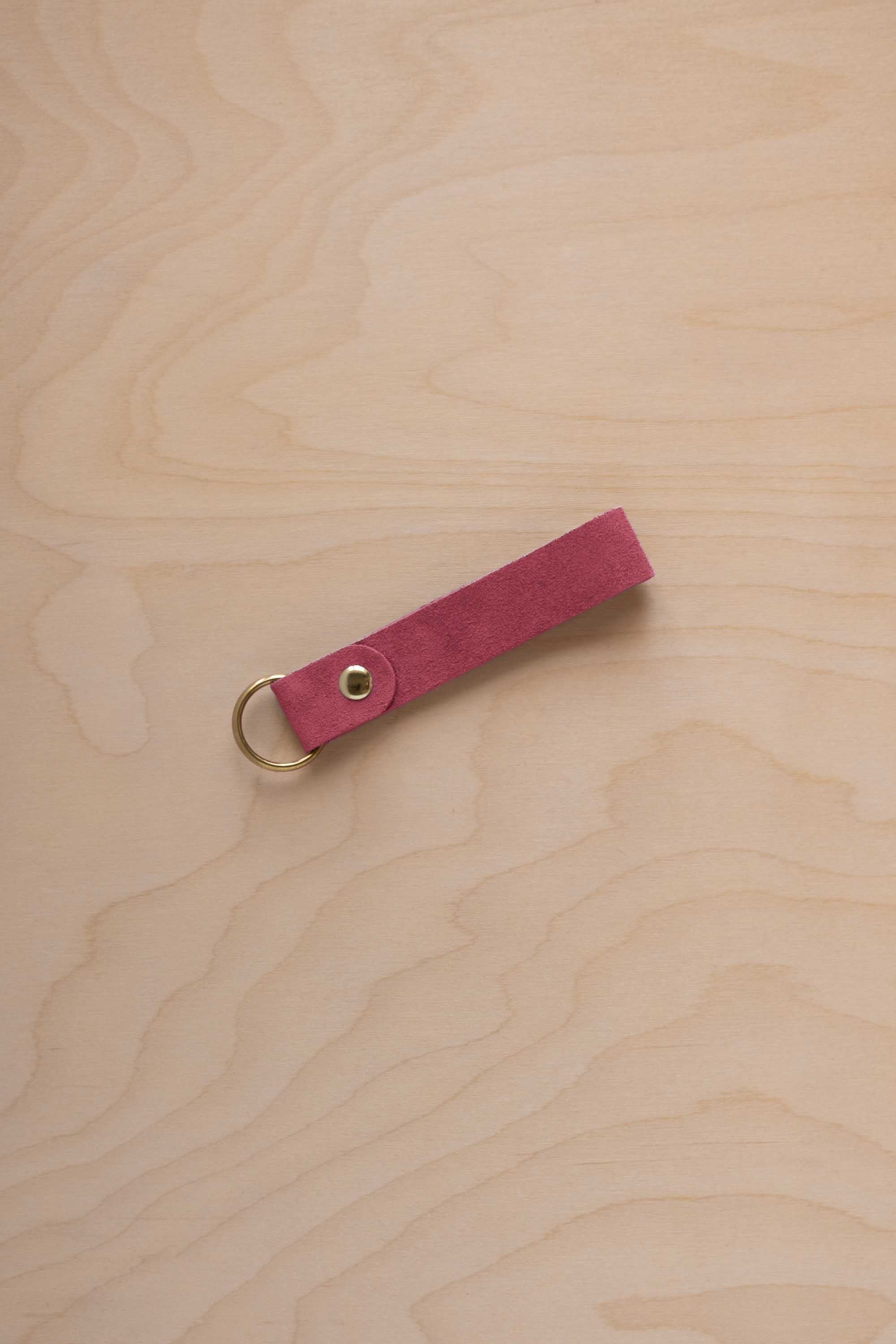 A faded rose pink coloured suede leather keychain with a gold metal ring and rivet, displayed diagonally on a light wood grain background.