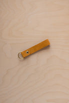 A golden ochre mustard yellow coloured beige suede leather keychain with a gold metal ring and rivet, displayed diagonally on a light wood grain background.