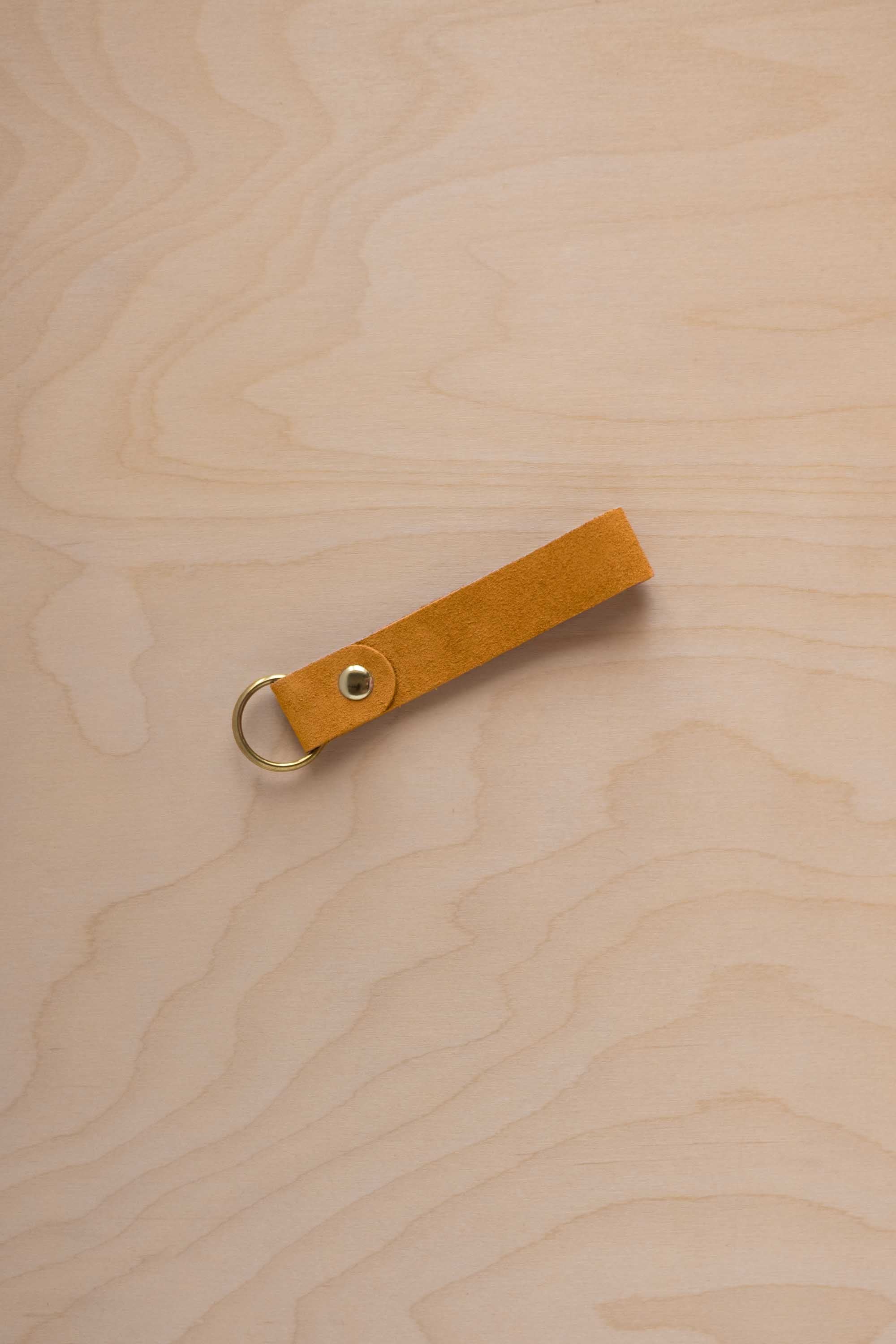 A golden ochre mustard yellow coloured beige suede leather keychain with a gold metal ring and rivet, displayed diagonally on a light wood grain background.