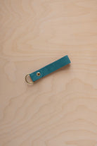 A marine teal coloured suede leather keychain with a gold metal ring and rivet, displayed diagonally on a light wood grain background.