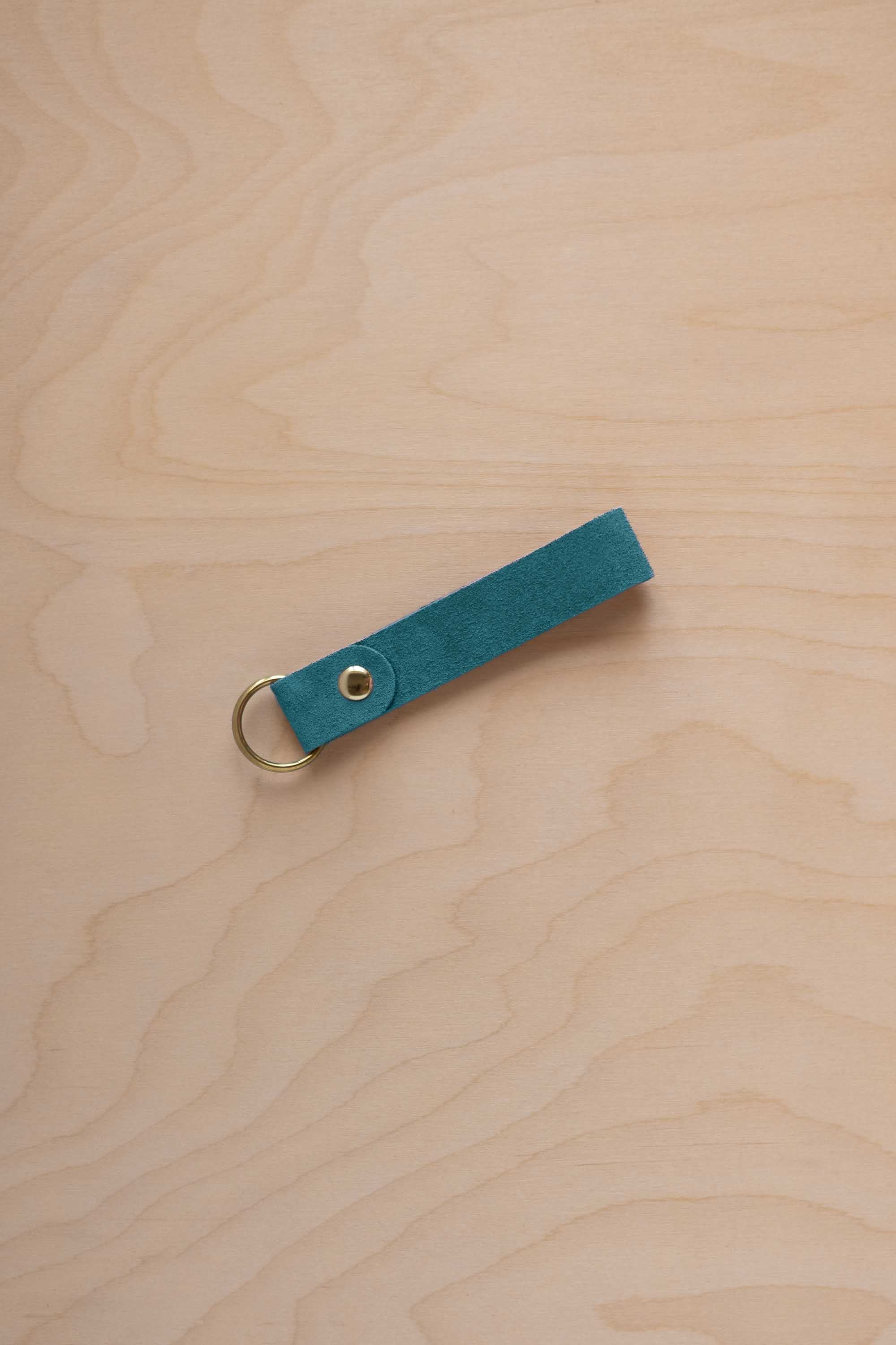 A marine teal coloured suede leather keychain with a gold metal ring and rivet, displayed diagonally on a light wood grain background.