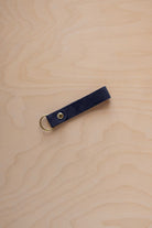 A midnight navy suede leather keychain with a gold metal ring and rivet, displayed diagonally on a light wood grain background.