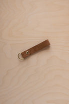 A mocha brown suede leather keychain with a gold metal ring and rivet, displayed diagonally on a light wood grain background.