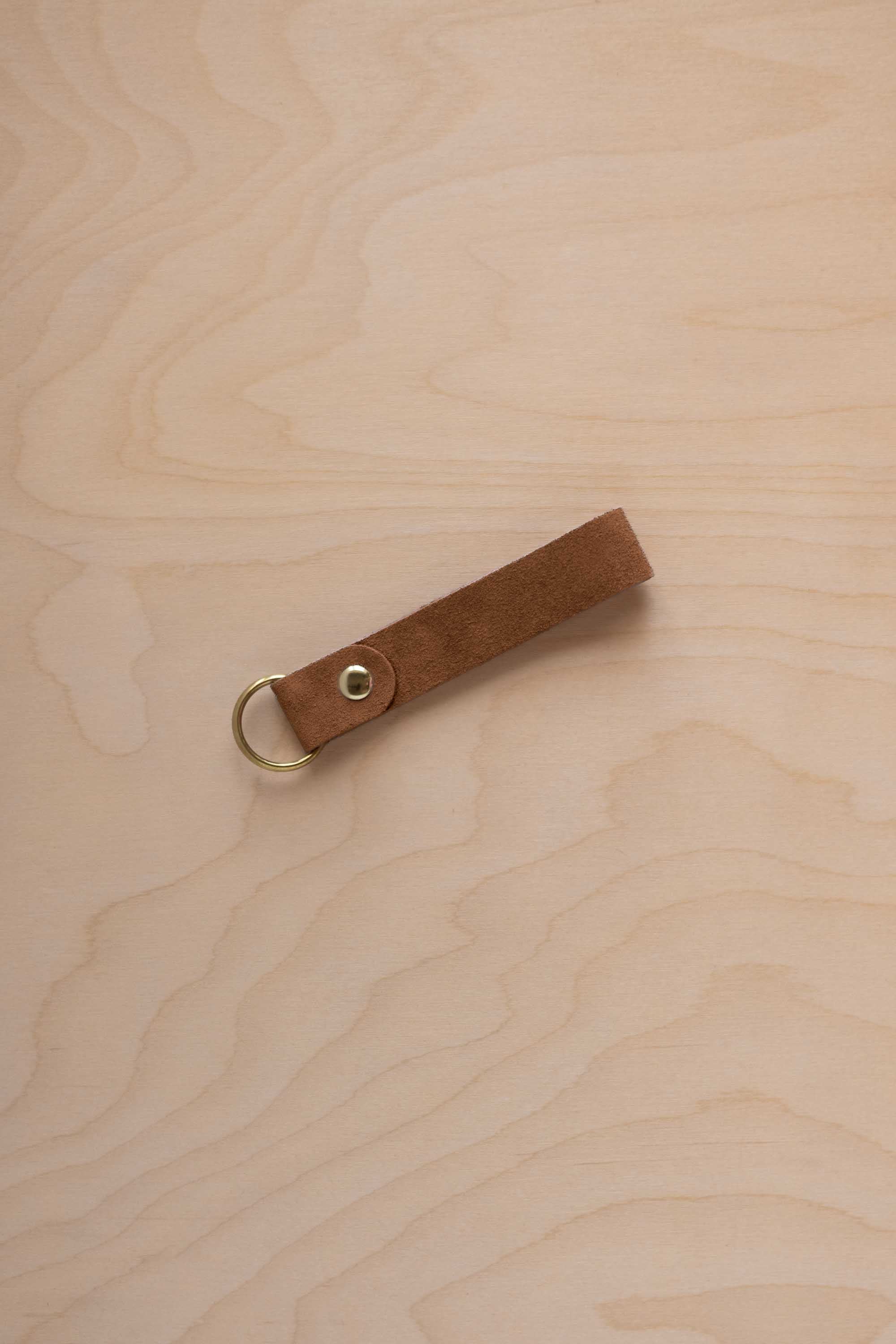 A mocha brown suede leather keychain with a gold metal ring and rivet, displayed diagonally on a light wood grain background.