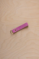 A peony pink suede leather keychain with a gold metal ring and rivet, displayed diagonally on a light wood grain background.