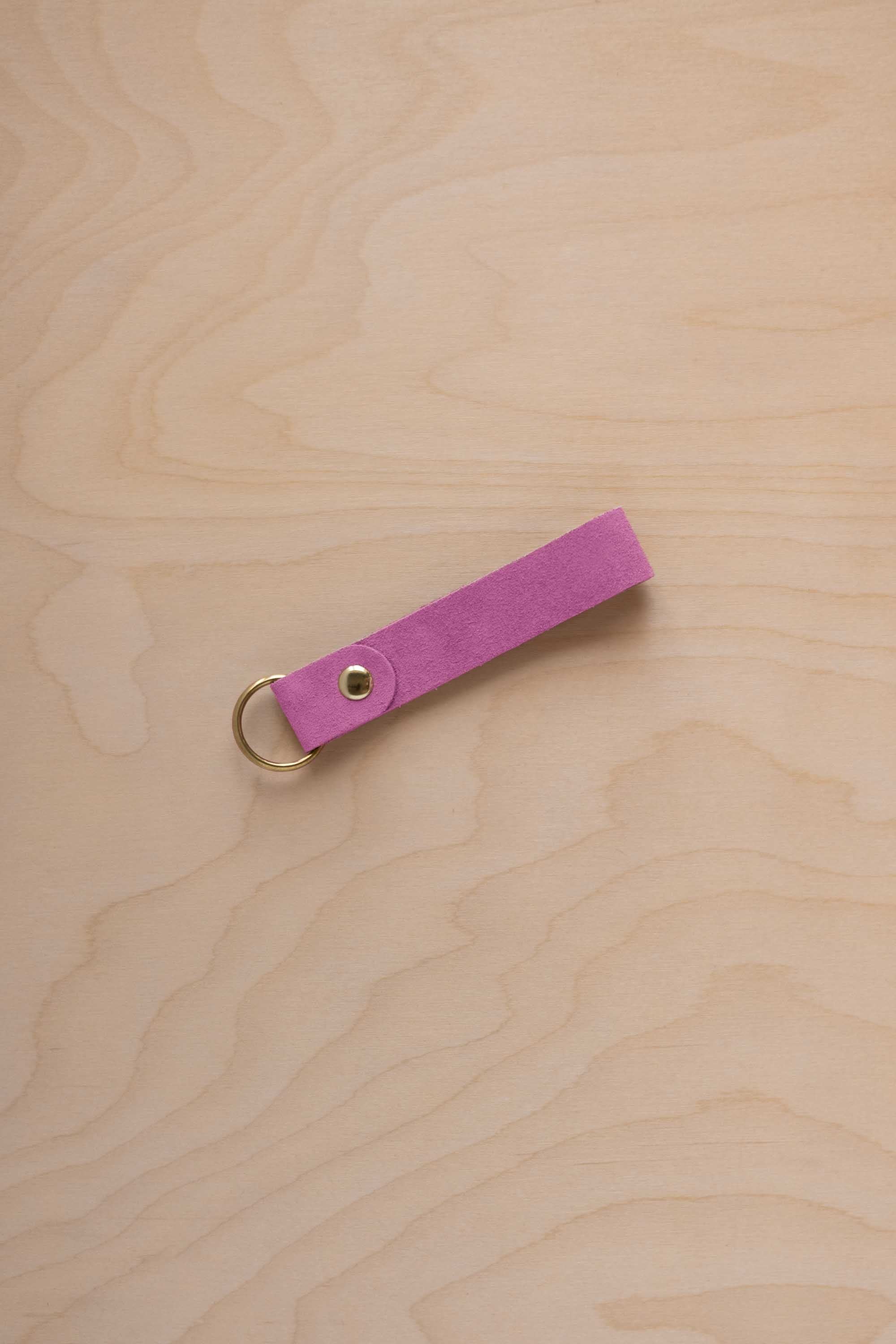 A peony pink suede leather keychain with a gold metal ring and rivet, displayed diagonally on a light wood grain background.