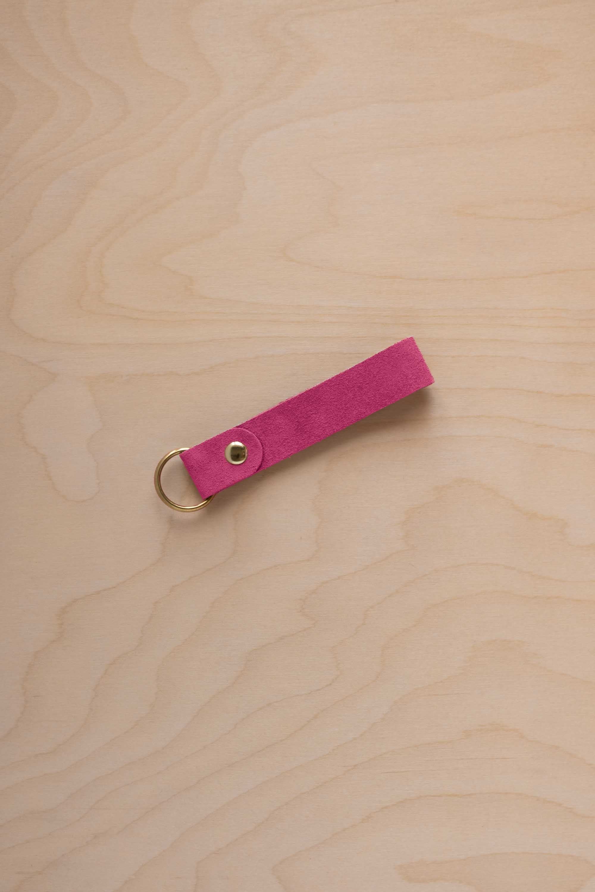 A raspberry pink suede leather keychain with a gold metal ring and rivet, displayed diagonally on a light wood grain background.