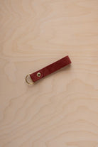A rust red suede leather keychain with a gold metal ring and rivet, displayed diagonally on a light wood grain background.