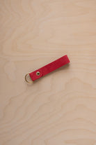 A scarlet red suede leather keychain with a gold metal ring and rivet, displayed diagonally on a light wood grain background.
