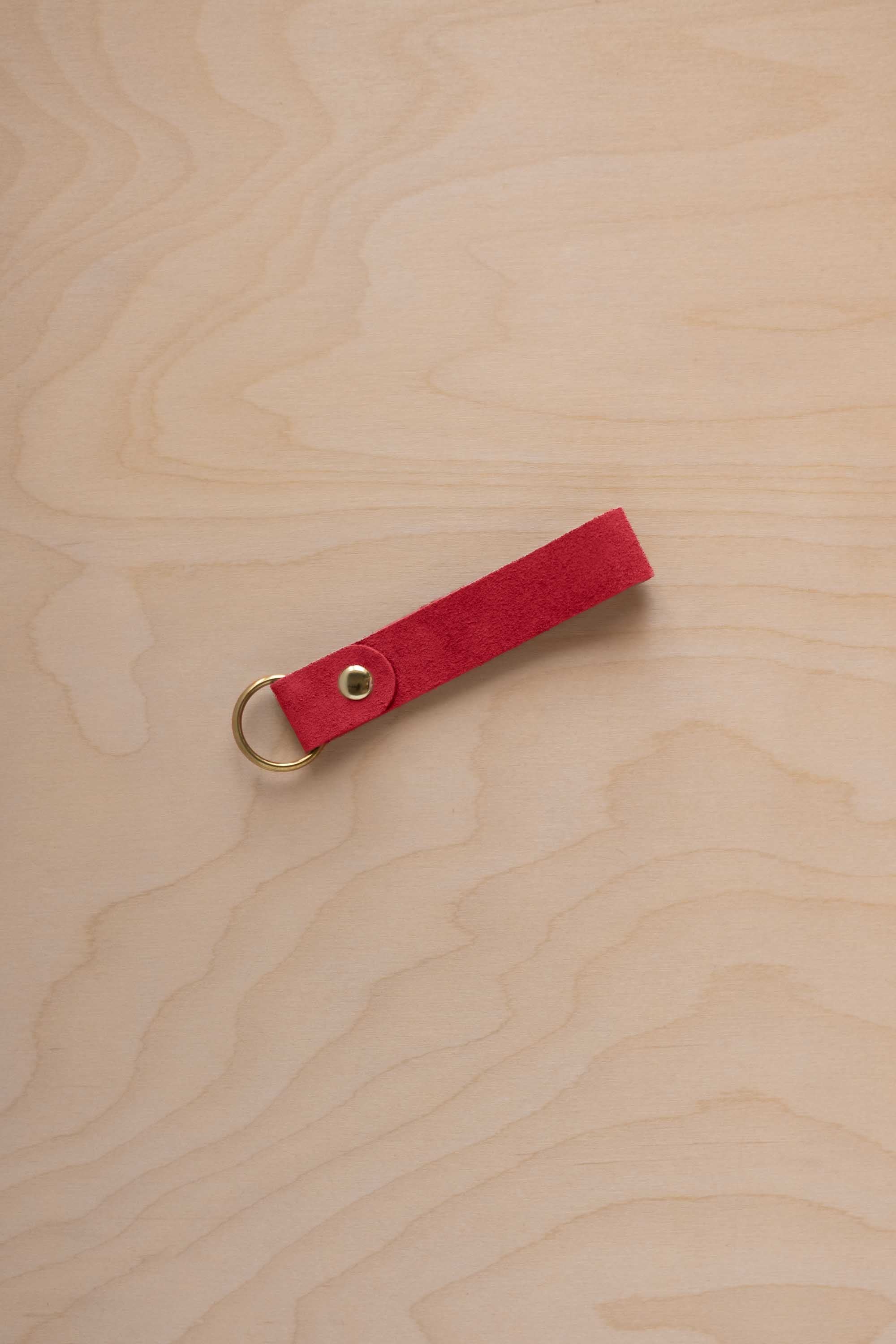 A scarlet red suede leather keychain with a gold metal ring and rivet, displayed diagonally on a light wood grain background.