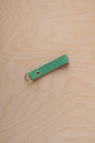 A seafoam turquoise green coloured suede leather keychain with a gold metal ring and rivet, displayed diagonally on a light wood grain background.