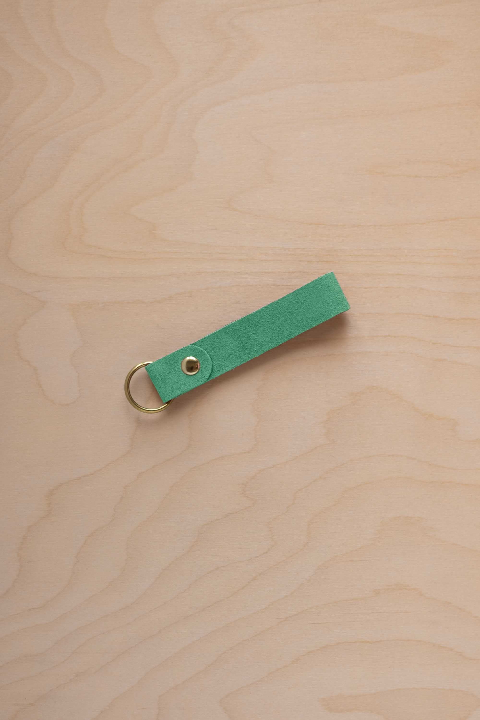 A seafoam turquoise green coloured suede leather keychain with a gold metal ring and rivet, displayed diagonally on a light wood grain background.