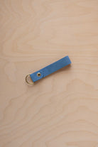 A sky blue suede leather keychain with a gold metal ring and rivet, displayed diagonally on a light wood grain background.