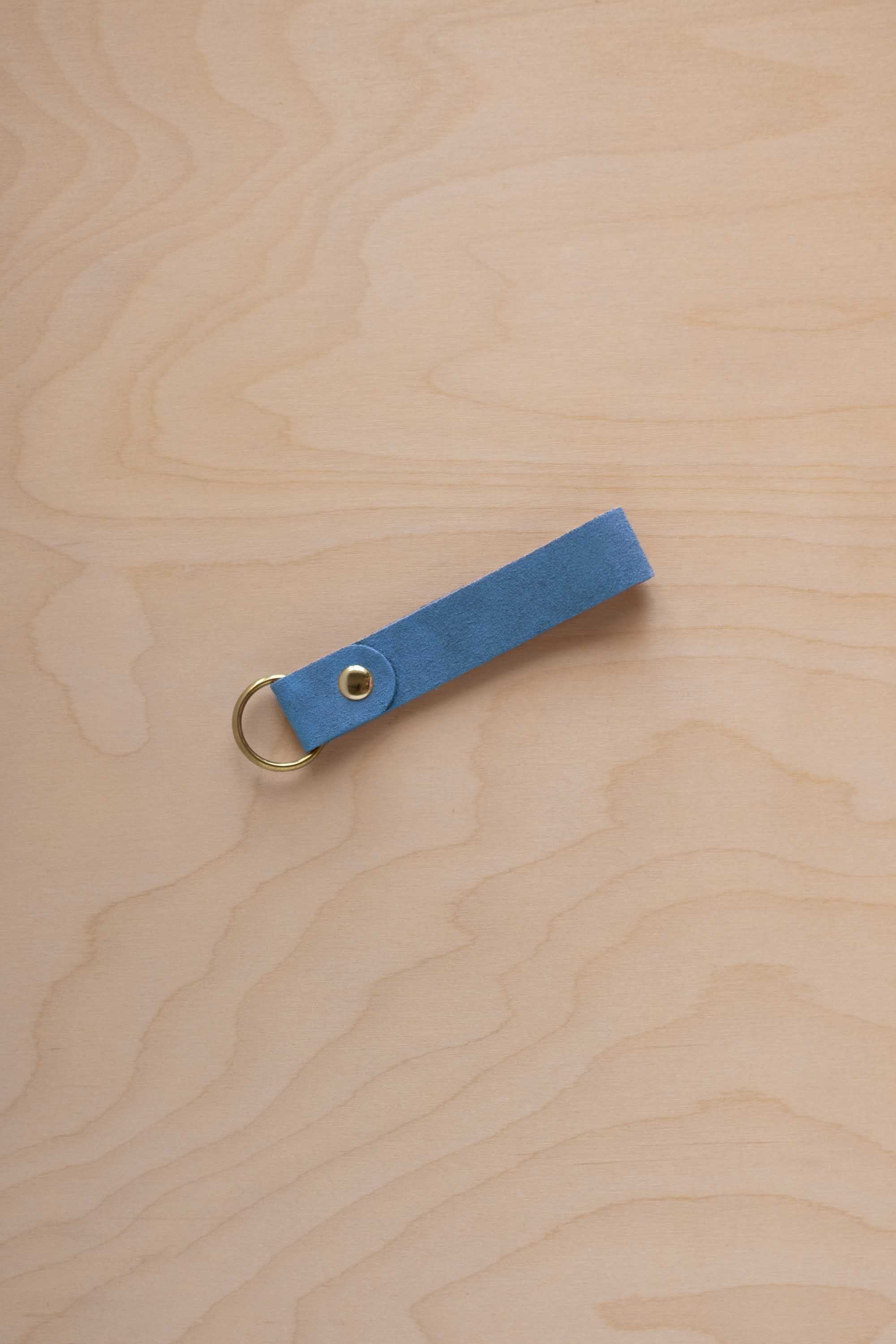 A sky blue suede leather keychain with a gold metal ring and rivet, displayed diagonally on a light wood grain background.