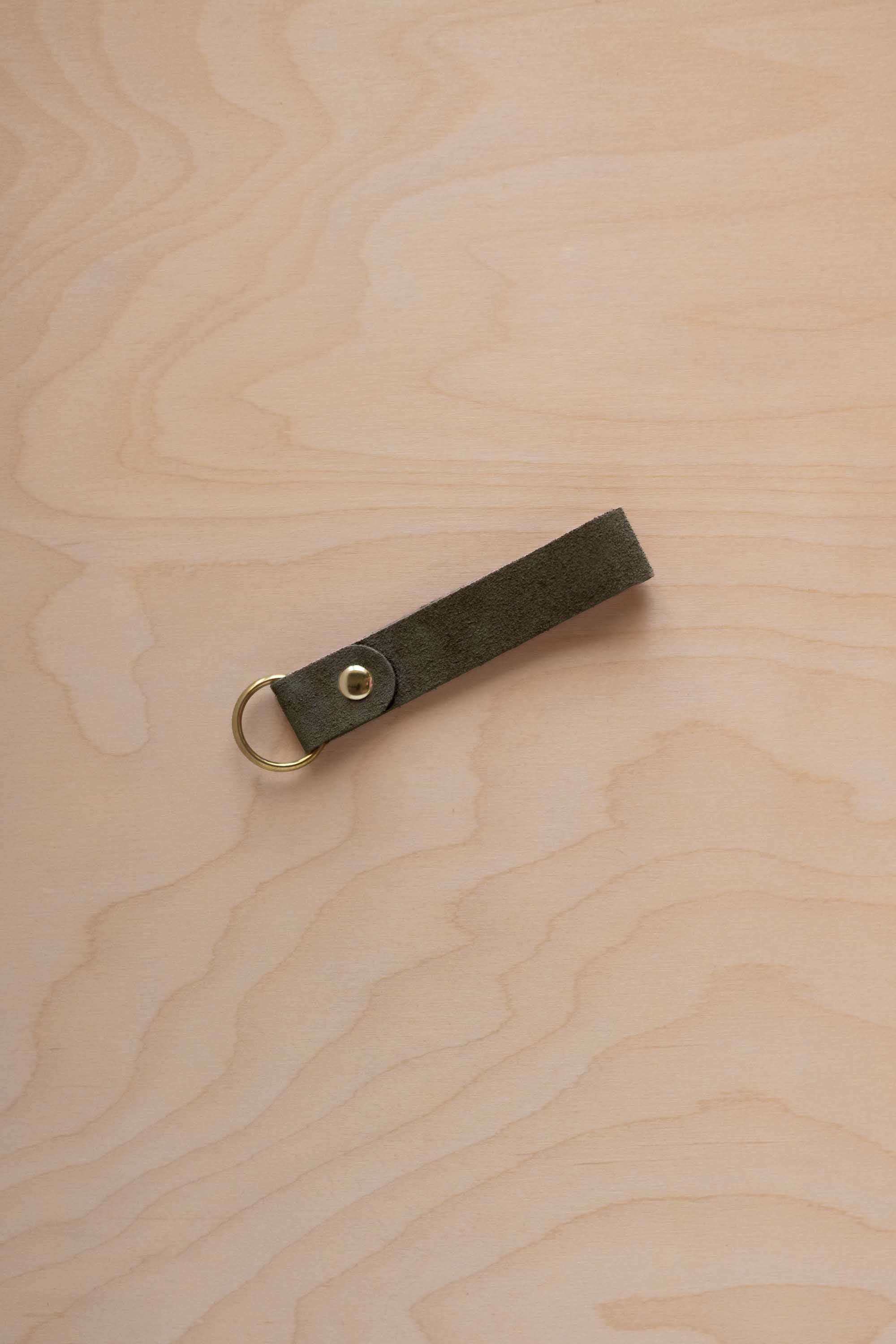 A soft olive green suede leather keychain with a gold metal ring and rivet, displayed diagonally on a light wood grain background.