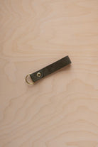 A soft olive green suede leather keychain with a gold metal ring and rivet, displayed diagonally on a light wood grain background.