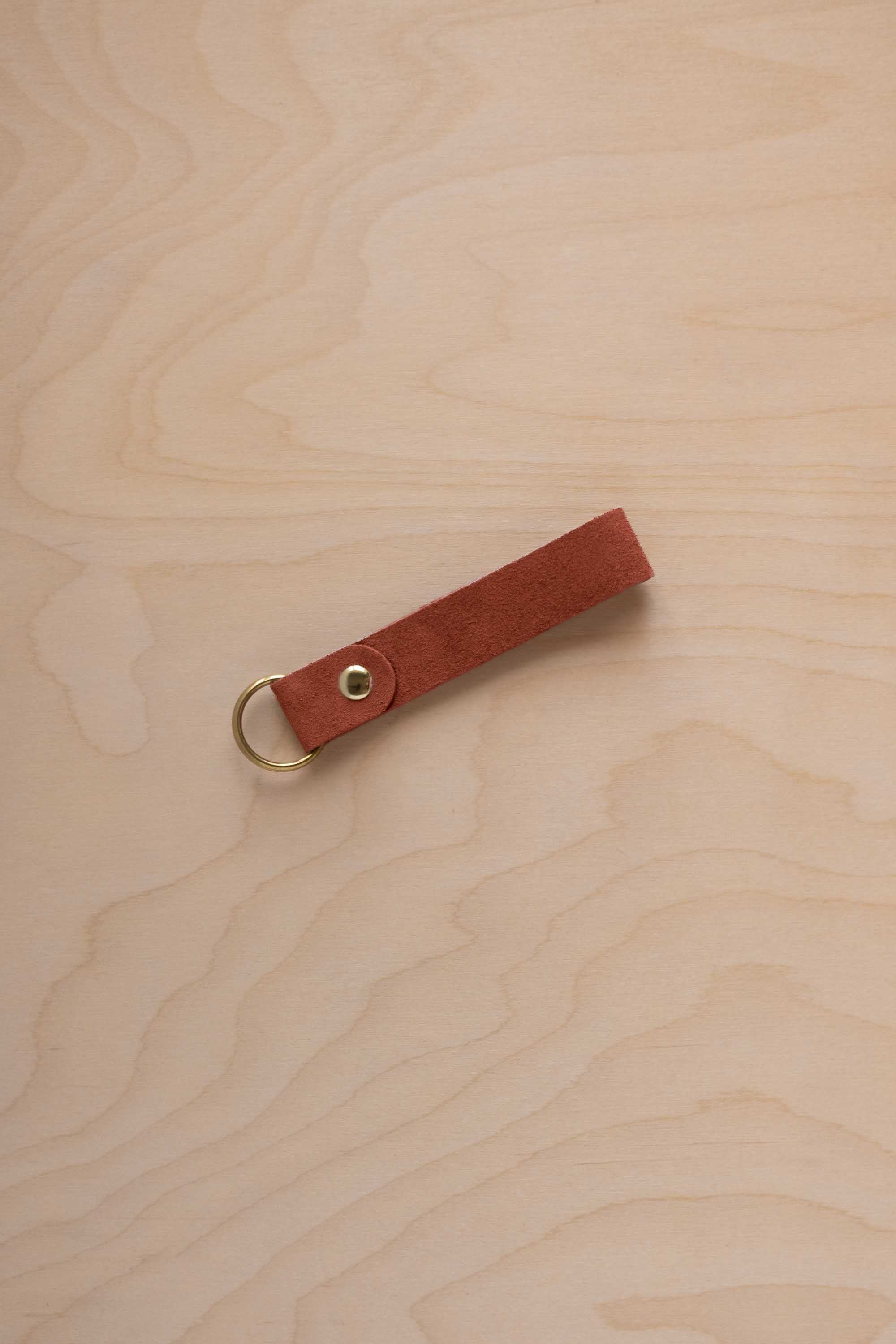 A terracotta suede leather keychain with a gold metal ring and rivet, displayed diagonally on a light wood grain background.