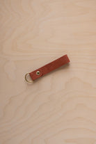 A terracotta suede leather keychain with a gold metal ring and rivet, displayed diagonally on a light wood grain background.