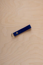 A ultramarine blue suede leather keychain with a gold metal ring and rivet, displayed diagonally on a light wood grain background.