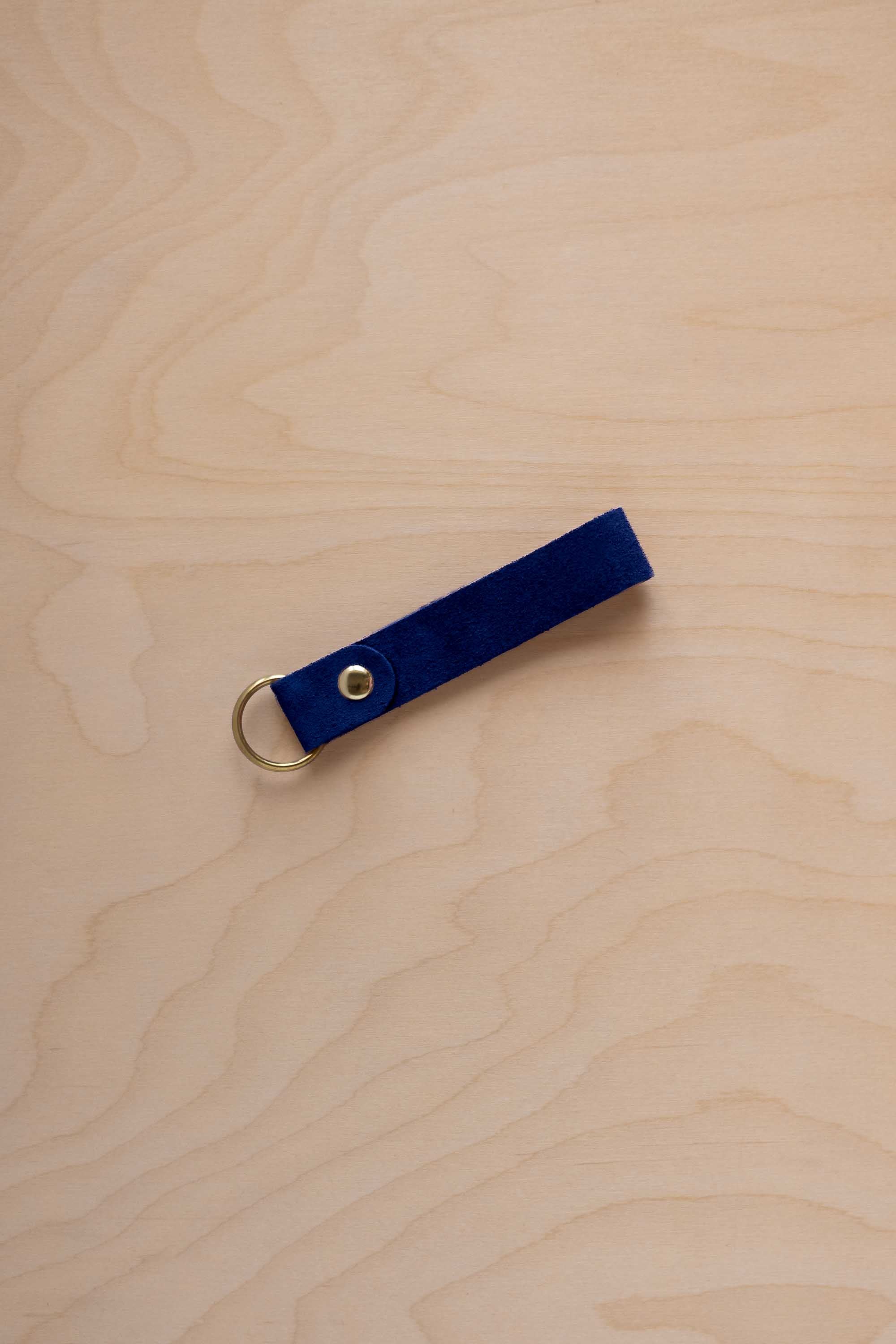 A ultramarine blue suede leather keychain with a gold metal ring and rivet, displayed diagonally on a light wood grain background.
